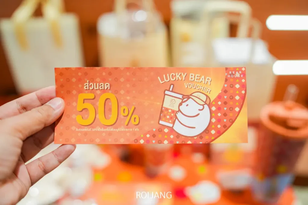 มือหนึ่งถือ "บัตรกำนัลหมีนำโชค" จาก BEARHOUSE มอบส่วนลด 50% พร้อมภาพประกอบหมีกำลังถือเครื่องดื่ม พื้นหลังสีส้มมีลวดลายละเอียดอ่อน.