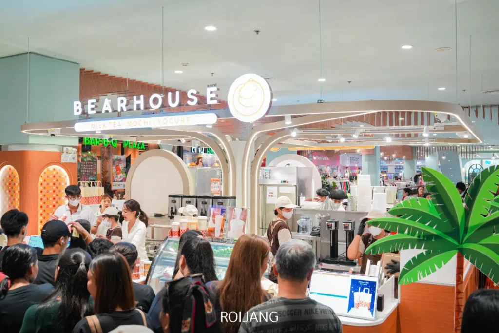 ผู้คนจำนวนมากยืนต่อแถวที่ BEARHOUSE ร้านชานมไข่มุกที่สว่างไสว พนักงานเตรียมเครื่องดื่มอย่างชำนาญอยู่หลังเคาน์เตอร์.