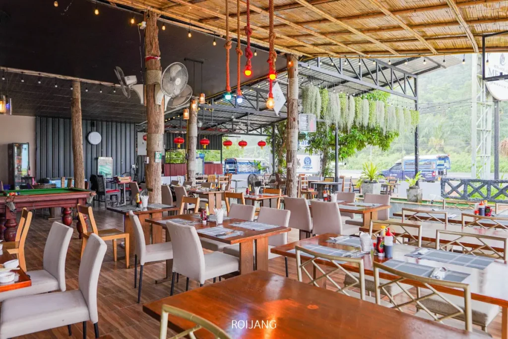 ร้านอาหารกลางแจ้ง Cynthia ที่เขาหลัก มีพื้นไม้ เก้าอี้สีอ่อน โต๊ะจัดวางอย่างเป็นระเบียบ ไฟประดับสาย และต้นไม้แขวน เต็มไปด้วยแสงธรรมชาติอ่อนๆ จากด้านข้าง.