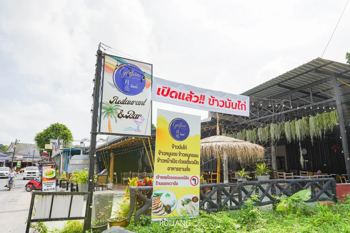 วิวภายนอกของร้านอาหาร Cynthia เขาหลัก พร้อมป้ายภาษาไทย รวมถึงป้ายแบนเนอร์ขนาดใหญ่ที่ประกาศการเปิดร้าน และป้ายเมนูอาหารที่ติดอยู่ใกล้ทางเข้า.