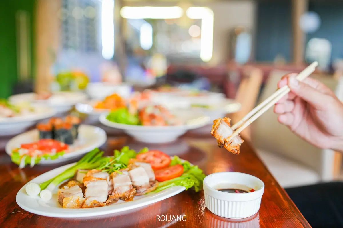 ที่ Cynthia เขาหลัก, คนใช้ตะเกียบหยิบชิ้นหมูสามชั้นย่างจากจาน ล้อมรอบด้วยอาหารหลากหลายชนิดและน้ำจิ้มบนโต๊ะอาหารในร้านอาหาร.