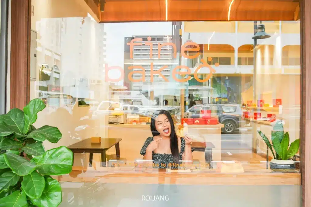 ผู้หญิงคนหนึ่งนั่งอยู่ที่โต๊ะภายในร้านเบเกอรี่ BEARHOUSE ยิ้มและถือขนมอบอยู่ เห็นผ่านหน้าต่างบานใหญ่ที่มีคำว่า "fine! bake" เขียนอยู่.