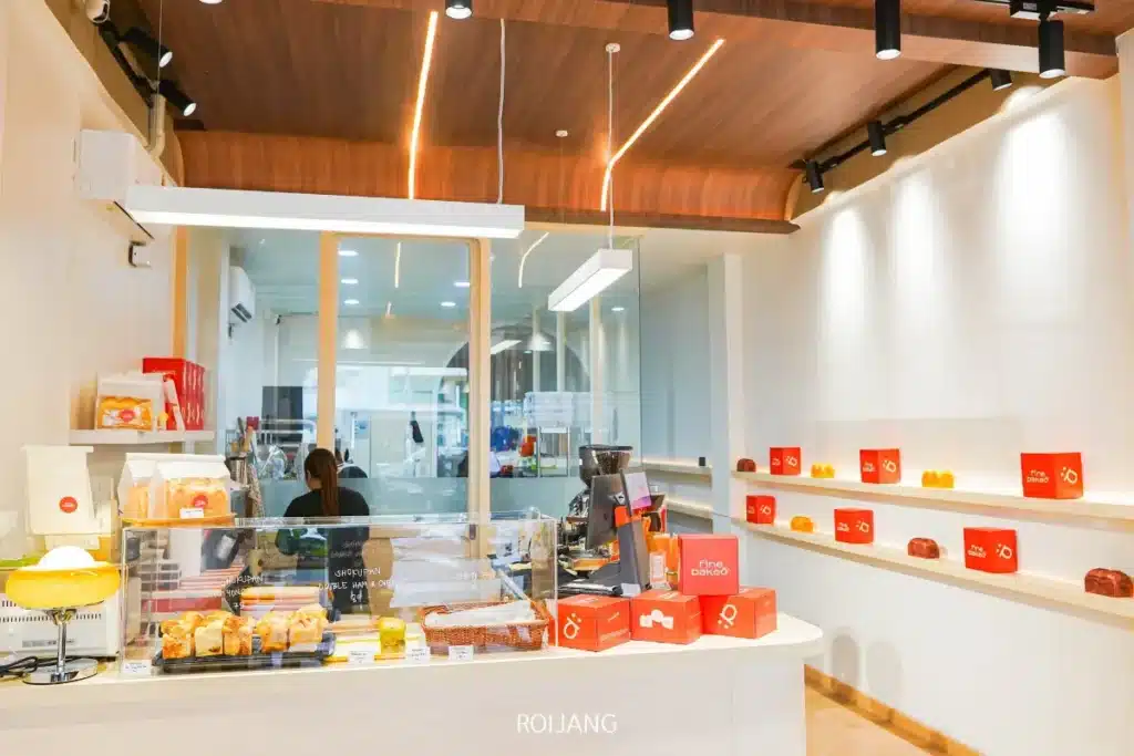 ภายในร้านเบเกอรี่ BEARHOUSE ทันสมัย มีเคาน์เตอร์แสดงขนมอบ บาร์ิสต้าทำงานอยู่หลังกระจก และชั้นวางกล่องสีแดงและขนมอบบนผนังสีขาว.