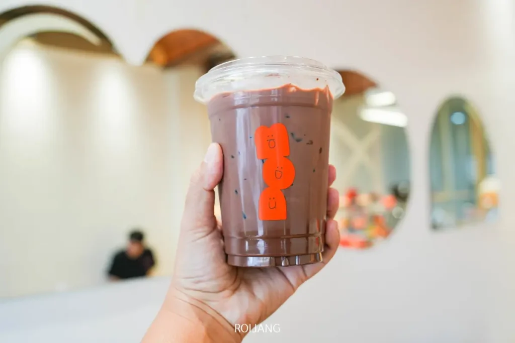 มือหนึ่งถือแก้วพลาสติกใส่น้ำแข็งช็อกโกแลตร้อนพร้อมฝาใสและมีโลโก้ BEARHOUSE สีส้ม อยู่ในบรรยากาศคาเฟ่สมัยใหม่ที่มีกระจกโค้งประดับผนัง.