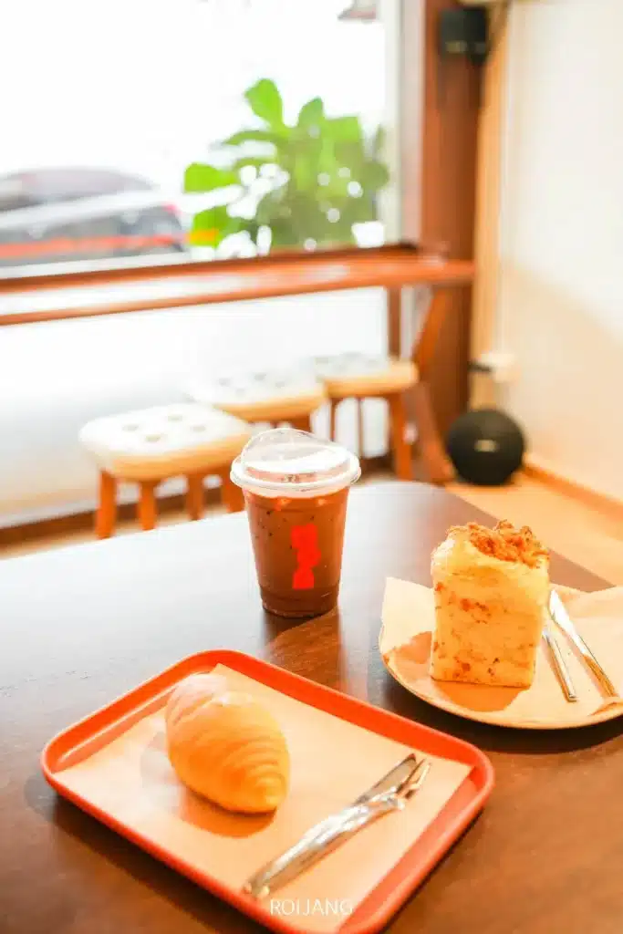 แก้วกาแฟเย็น ครัวซองต์ และขนมปังรสเค็มหนึ่งชิ้นวางอยู่บนถาดที่โต๊ะไม้ในร้าน BEARHOUSE cafe โดยมีเก้าอี้สตูลและหน้าต่างเป็นฉากหลัง.