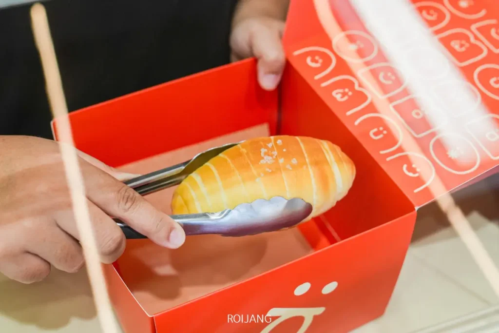 บุคคลหนึ่งวางขนมปังโรลลงในกล่อง BEARHOUSE สีแดงโดยใช้คีมโลหะ.