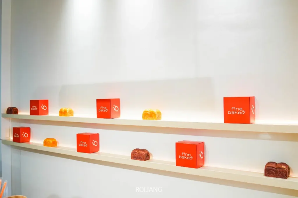 ชั้นวางของติดผนังสีขาวสองชั้นแสดงกล่อง BEARHOUSE "fine bake6" สีส้มสลับกันและขนมอบหลากหลายชนิดภายใต้แสงไฟที่สว่างจ้า.