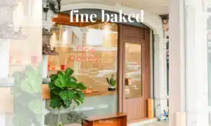 มุมมองถนนของร้านเบเกอรี่ BEARHOUSE ที่มีประตูไม้ หน้าต่างบานใหญ่แสดงคำว่า "Fine Baked" พืชสีเขียวอยู่ด้านนอก และหน้าร้านที่สว่างสดใสและเชิญชวน.