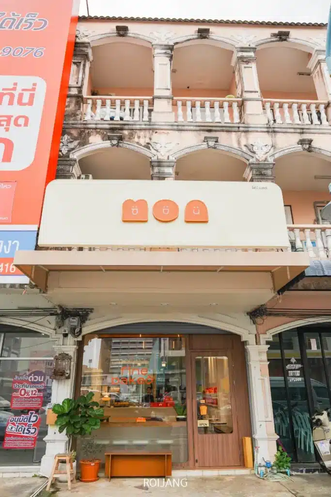ร้านเบเกอรี่ขนาดเล็กชื่อ BEARHOUSE มีป้ายสีเบจและทางเข้าเป็นกระจก ตั้งอยู่บนชั้นหนึ่งของอาคารสามชั้นที่ดูเก่าและมีระเบียง.