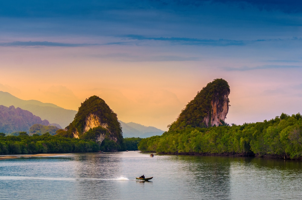เรือลำเล็กแล่นอยู่บนผืนน้ำที่สงบ โดยมีภูเขาหินปูนขนาดใหญ่สองลูกที่ปกคลุมไปด้วยป่าเขียวขจีเป็นฉากหลัง ท่ามกลางท้องฟ้ายามพระอาทิตย์ตกที่เต็มไปด้วยสีสัน—ภาพอันเงียบสงบที่ถ่ายทอดความงดงามของสถานที่ท่องเที่ยวกระบี่.