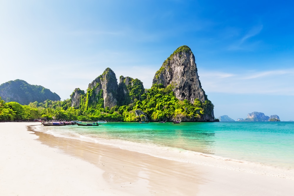 ชายหาดเขตร้อนที่มีน้ำทะเลใสสีฟ้าเทอร์ควอยซ์ ทรายขาว ต้นไม้เขียวขจี และหน้าผาหินปูนสูงตระหง่านใต้ท้องฟ้าสีฟ้าสดใส—เหมาะสำหรับผู้ที่ต้องการ เที่ยวกระบี่ และสัมผัสความงดงามตามธรรมชาติของ สถานที่ท่องเที่ยวในกระบี่.