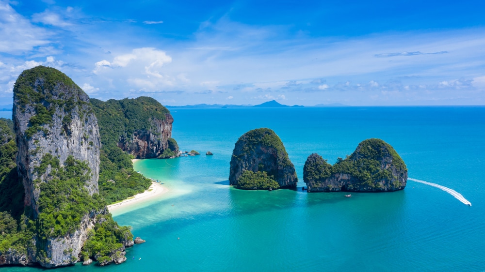 ภาพมุมสูงของทะเลสีเทอร์ควอยซ์ ชายหาดทรายขาว และหน้าผาหินปูนที่มีเกาะเล็ก ๆ อยู่เบื้องหน้า—สถานที่ท่องเที่ยวที่น่าตื่นตาตื่นใจในกระบี่—พร้อมเรือเร็วที่แล่นผ่านน้ำใต้ท้องฟ้าสีคราม.