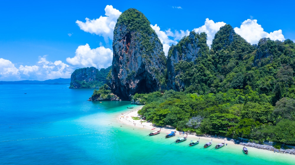 ภาพมุมสูงของชายหาดเขตร้อนในสถานที่ท่องเที่ยวกระบี่ มีน้ำทะเลสีฟ้าเทอร์ควอยซ์ ทรายขาว เรือที่จอดทอดสมอ และหน้าผาหินปูนสูงใหญ่ปกคลุมด้วยพืชพรรณสีเขียว ท่ามกลางท้องฟ้าสีครามและเมฆลอย.