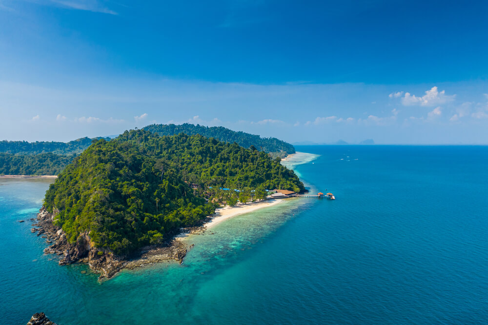 ภาพมุมสูงของเกาะเขียวชอุ่มที่ล้อมรอบด้วยมหาสมุทรสีฟ้าใส มีชายหาดทรายและท่าเทียบเรือเล็กๆ เป็นสถานที่ท่องเที่ยวในกระบี่ที่เชิญชวน เหมาะสำหรับผู้ที่ฝันถึงการเดินทางในกระบี่และพักผ่อนท่ามกลางความงามตามธรรมชาติ.