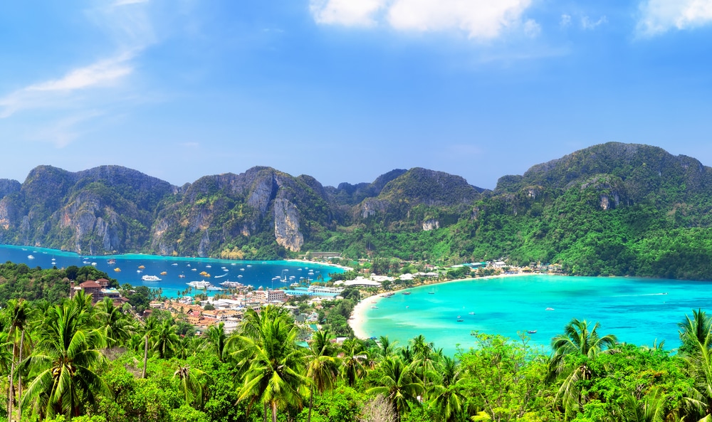 ภาพมุมสูงของชายฝั่งเขตร้อนที่มีน้ำสีฟ้าเทอร์ควอยซ์ ชายหาดทรายสีขาว ภูเขาเขียวขจี ต้นปาล์ม และเรือที่จอดอยู่ใกล้เมืองชายฝั่ง—เหมาะสำหรับผู้ที่ต้องการเที่ยวกระบี่หรือค้นหาสถานที่ท่องเที่ยวใหม่ในกระบี่.