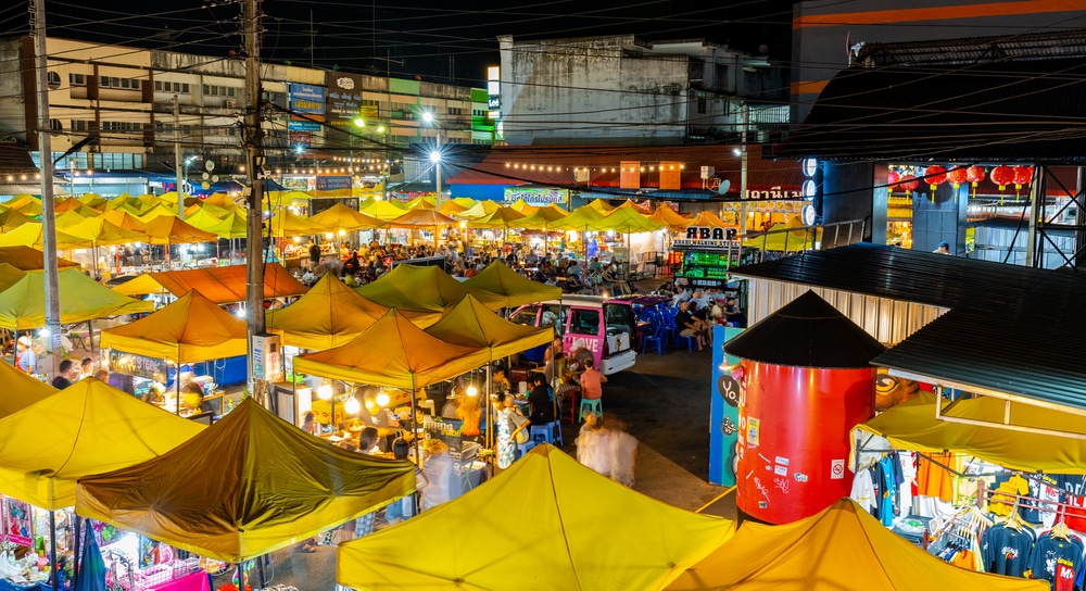 ภาพยามค่ำคืนของตลาดกลางแจ้งที่มีเต็นท์สีเหลือง ไฟสว่างไสว แผงขายของมากมาย และผู้คนกำลังจับจ่ายซื้อของ—บรรยากาศคึกคักเหมาะสำหรับผู้ที่กำลังมองหา ที่เที่ยวกระบี่ โดยมีอาคารและสายไฟปรากฏให้เห็นอยู่เบื้องหลัง.