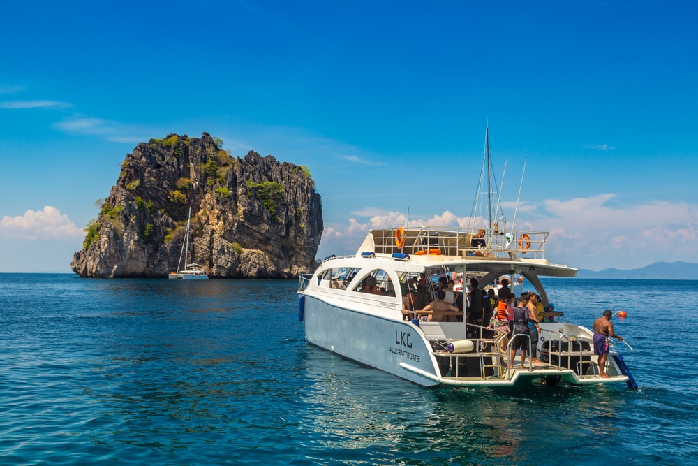 เรือสีขาวลำหนึ่งที่มีผู้คนอยู่บนเรือลอยอยู่ใกล้เกาะหิน ในขณะที่เรือใบอีกลำหนึ่งจอดทอดสมออยู่ใกล้ๆ ภายใต้ท้องฟ้าสีฟ้าใส—ภาพที่สมบูรณ์แบบสำหรับใครก็ตามที่กำลังมองหา เที่ยวกระบี่ หรือค้นพบ สถานที่ท่องเที่ยวใหม่ในกระบี่.