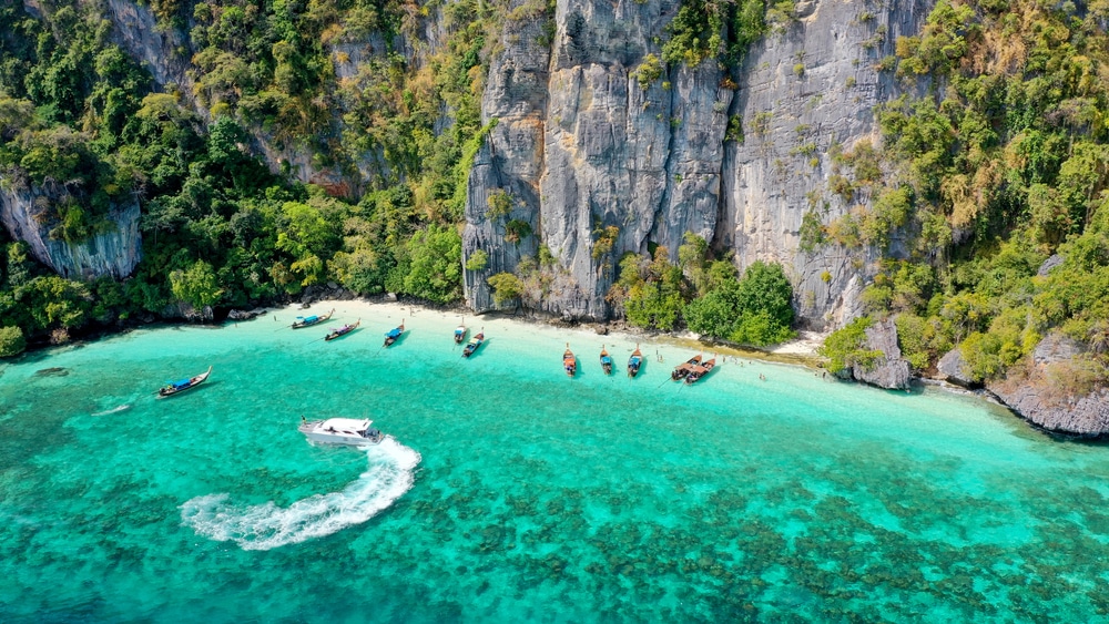 ภาพมุมสูงของน้ำสีฟ้าเทอร์ควอยซ์ เรือสีขาวแล่นทิ้งรอยคลื่น เรือลำเล็กหลายลำจอดเรียงรายใกล้ชายหาดทราย และหน้าผาสูงชันปกคลุมด้วยพืชพรรณเขียวขจี—เหมาะสำหรับผู้ที่ต้องการเที่ยวกระบี่และค้นพบสถานที่ลับอันน่าหลงใหล.