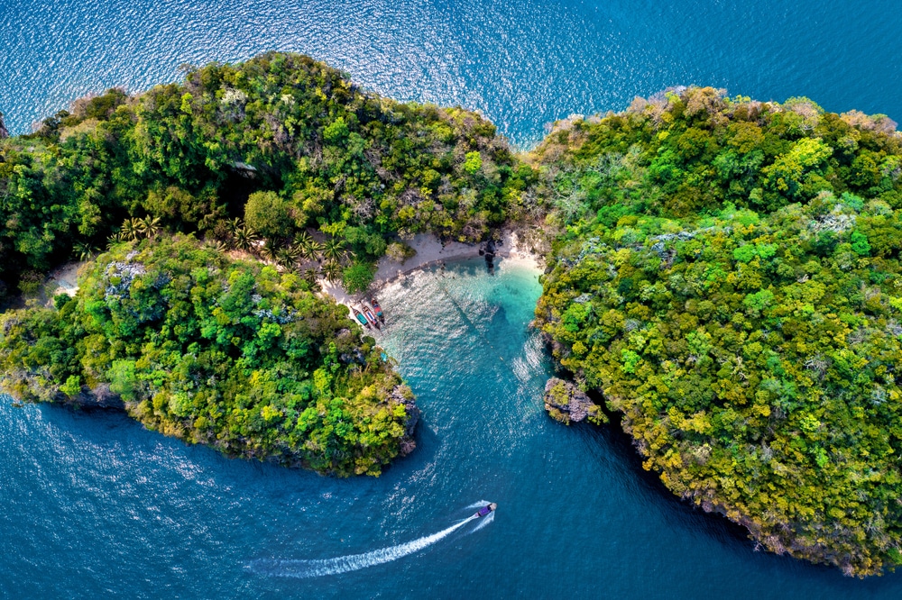 ภาพมุมสูงของเกาะสองเกาะที่เขียวชอุ่มและอุดมสมบูรณ์ แยกจากกันด้วยน้ำทางแคบ ๆ ในที่เที่ยวกระบี่ พร้อมเรือเล็ก ๆ ที่ทิ้งรอยคลื่นสีขาวในน้ำสีคราม.