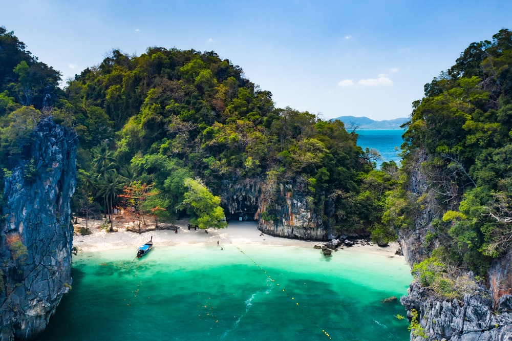 ชายหาดเขตร้อนที่เงียบสงบในสถานที่ท่องเที่ยวกระบี่ มีน้ำทะเลใสสีเทอร์ควอยซ์ ล้อมรอบด้วยหน้าผาหินและต้นไม้เขียวชอุ่ม มีเรือลำเล็กจอดทอดสมออยู่ใกล้ชายฝั่ง.