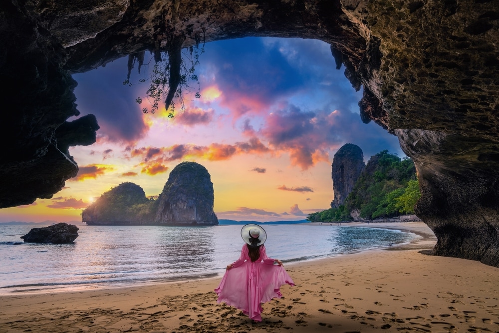 บุคคลในชุดเดรสสีชมพูและหมวกปีกกว้างยืนอยู่บนชายหาดทรายใต้ถ้ำหิน มองไปยังทะเลและเกาะหินปูนที่ห่างไกลในยามพระอาทิตย์ตกดิน—ช่วงเวลาที่น่าจดจำไม่รู้ลืมขณะเที่ยวกระบี่.