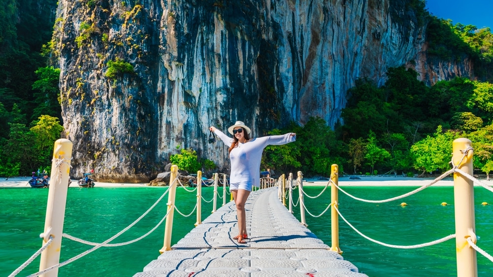 ผู้หญิงในชุดลำลองและหมวกยืนกางแขนออกบนท่าเรือลอยน้ำเหนือผืนน้ำสีฟ้าคราม มีหน้าผาหินสูงและพืชพรรณเขียวชอุ่มเป็นฉากหลัง—ช่วงเวลาที่สมบูรณ์แบบสำหรับผู้ที่กำลังมองหา เที่ยวกระบี่.
