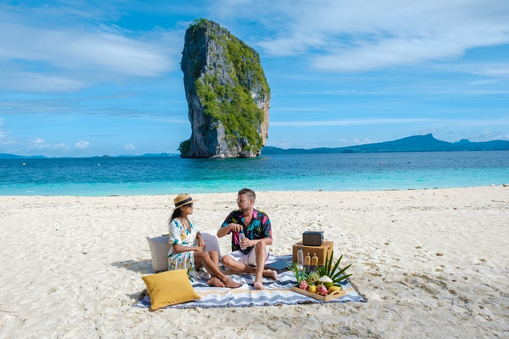 สองคนกำลังปิกนิกบนชายหาดทรายที่มีน้ำทะเลใสสีฟ้าและเกาะหินสูงเป็นฉากหลัง; เหมาะสำหรับใครก็ตามที่กำลังมองหา ที่เที่ยวกระบี่, อุปกรณ์ปิกนิก และวิทยุถูกวางไว้บนผ้าปู.