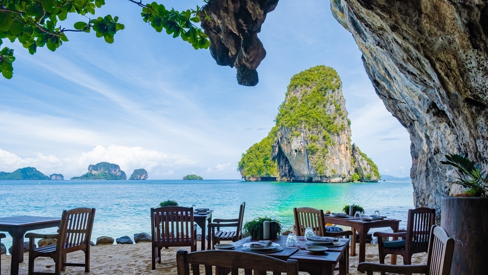 ร้านอาหารกลางแจ้งบนชายหาดทราย มีโต๊ะและเก้าอี้ไม้ ตั้งอยู่ใต้หน้าผาหิน มองเห็นน้ำทะเลสีฟ้าครามและเกาะหินปูนขนาดใหญ่ เหมาะสำหรับผู้ที่ต้องการ เที่ยวกระบี่.