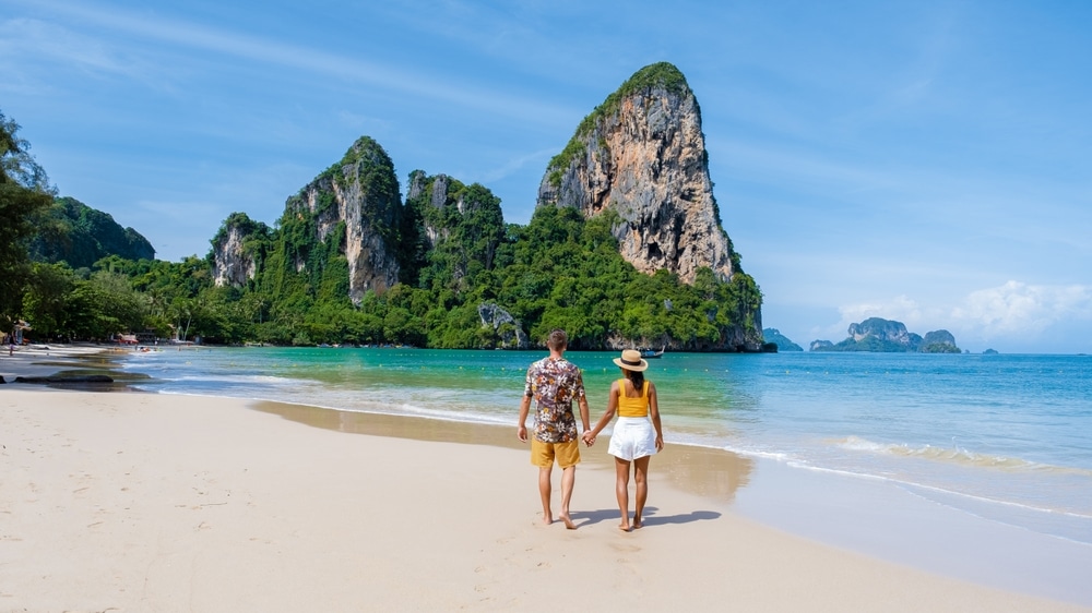 คู่รักคู่หนึ่งเดินเล่นบนชายหาดทรายขาวสะอาด ทะเลสีฟ้าใส ภูเขาหินเขียวชอุ่ม และยอดเขาหินสูงตระหง่านเป็นฉากหลัง ภายใต้ท้องฟ้าสดใส—เหมาะสำหรับผู้ที่ต้องการไปเที่ยวกระบี่ ท่ามกลางทัศนียภาพที่สวยงามตระการตา.
