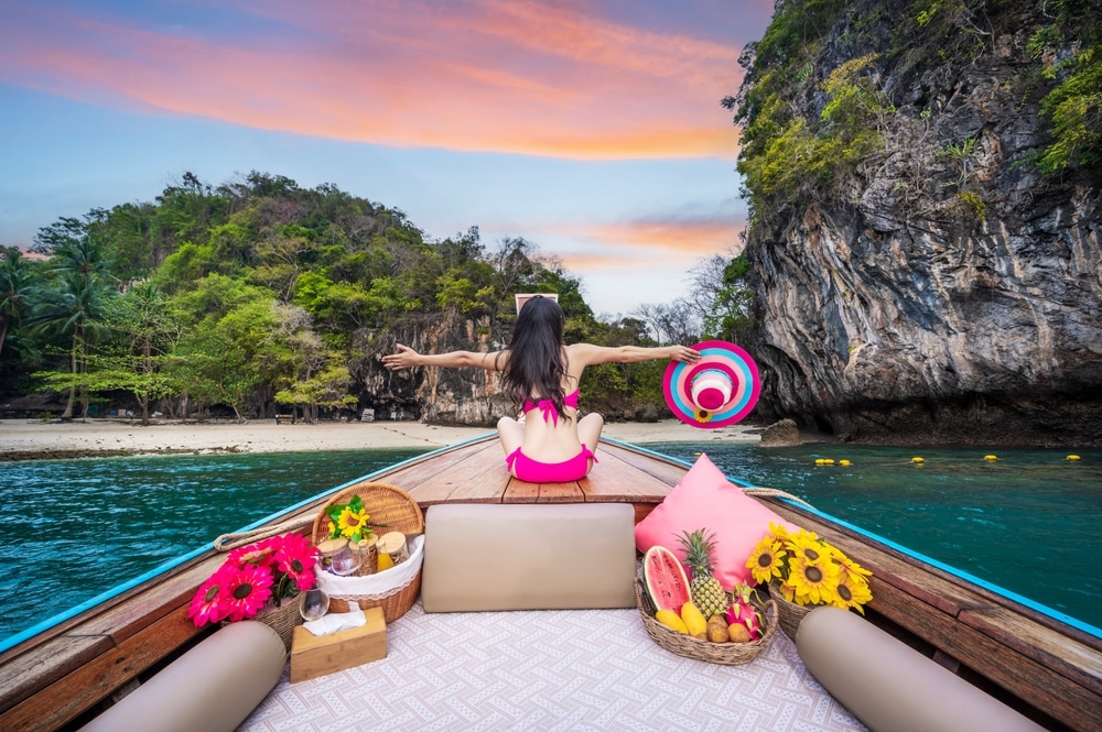 บุคคลในชุดว่ายน้ำสีชมพู นั่งอยู่บนเรือ หันหน้าไปทางเกาะ แขนทั้งสองข้างยื่นออกไป หมวกสีสันสดใสอยู่ในมือ มีผลไม้และดอกไม้บนดาดฟ้าในยามพระอาทิตย์ตก—ถ่ายทอดความมหัศจรรย์ของ ที่เที่ยวกระบี่.