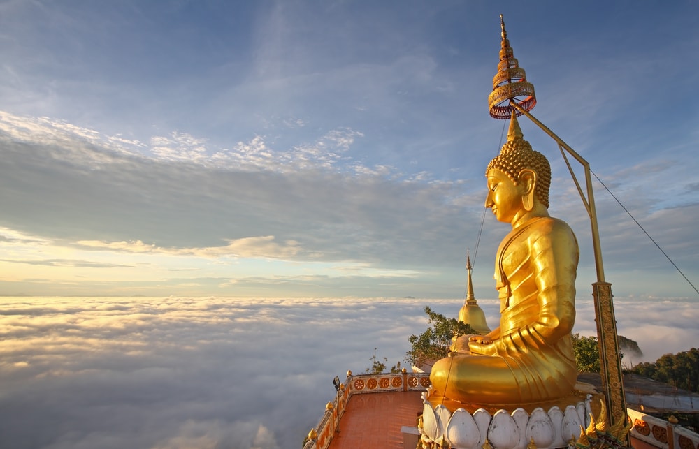 พระพุทธรูปทองคำขนาดใหญ่ตั้งตระหง่านเหนือทะเลหมอกและภูเขาไกลโพ้นในยามพระอาทิตย์ขึ้นหรือพระอาทิตย์ตกดิน พร้อมด้วยโครงสร้างของวัดที่ตั้งอยู่ฐานของพระพุทธรูป—ภาพที่น่าตื่นตาตื่นใจสำหรับผู้ที่มาเยือนสถานที่ท่องเที่ยวในกระบี่.