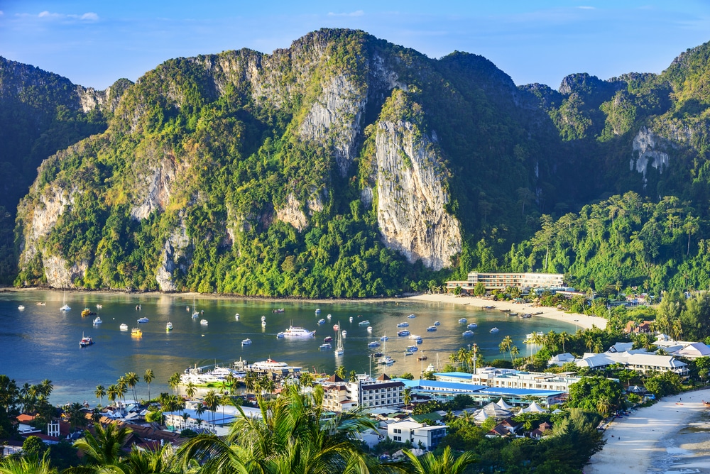 เมืองชายฝั่งที่มีอาคารสีขาวและเรือในอ่าว ล้อมรอบด้วยหน้าผาและภูเขาที่ปกคลุมไปด้วยสีเขียวภายใต้ท้องฟ้าสีคราม—เหมาะสำหรับผู้ที่กำลังมองหาสถานที่ท่องเที่ยวในกระบี่สำหรับการเดินทางครั้งต่อไป.