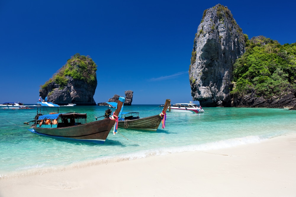 เรือไม้สองลำลอยอยู่บนน้ำสีฟ้าใสราวกับเทอร์ควอยซ์ ใกล้ชายหาดทรายและเกาะหินเขียวชอุ่ม ชวนให้คุณมาสัมผัสความงดงามอันน่าทึ่งของสถานที่ท่องเที่ยวในกระบี่ใต้ท้องฟ้าสีครามสดใส.