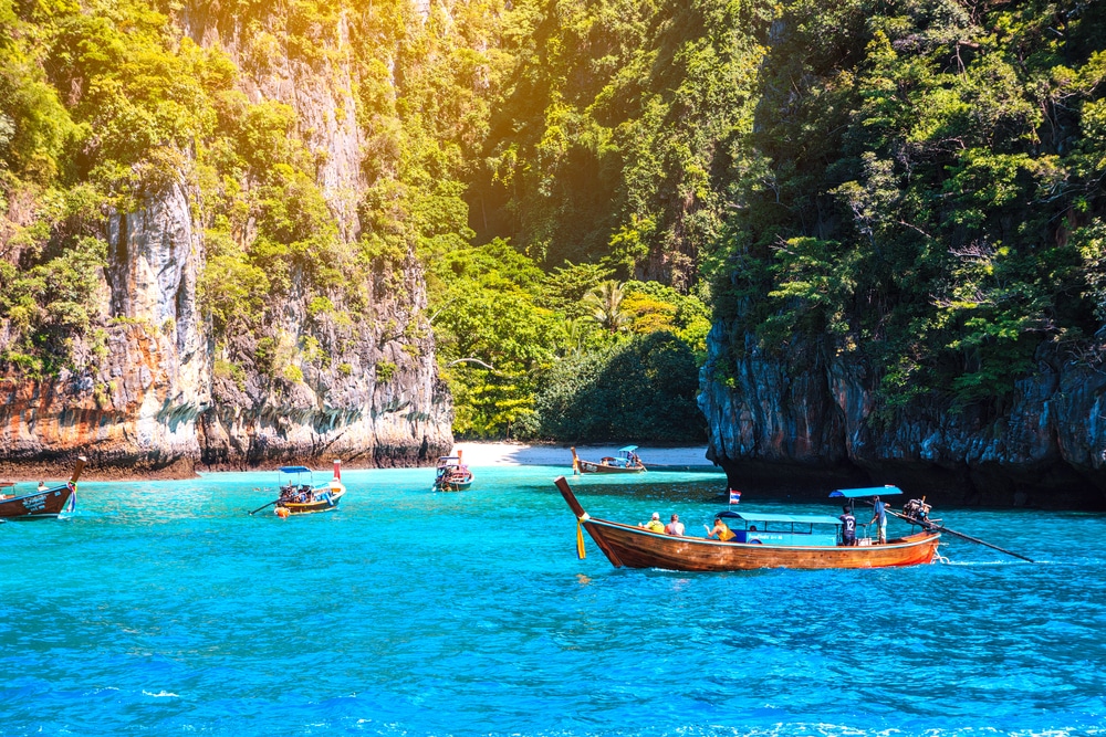 เรือไม้ลอยอยู่บนน้ำสีฟ้าสดใสใกล้ชายฝั่งที่เต็มไปด้วยโขดหินและป่าไม้ มีหน้าผาสูงชันและพืชพรรณเขียวชอุ่ม—ภาพอันงดงามที่เหมาะสำหรับผู้ที่ต้องการสำรวจสถานที่ท่องเที่ยวในกระบี่หรือค้นพบความงามของกระบี่.