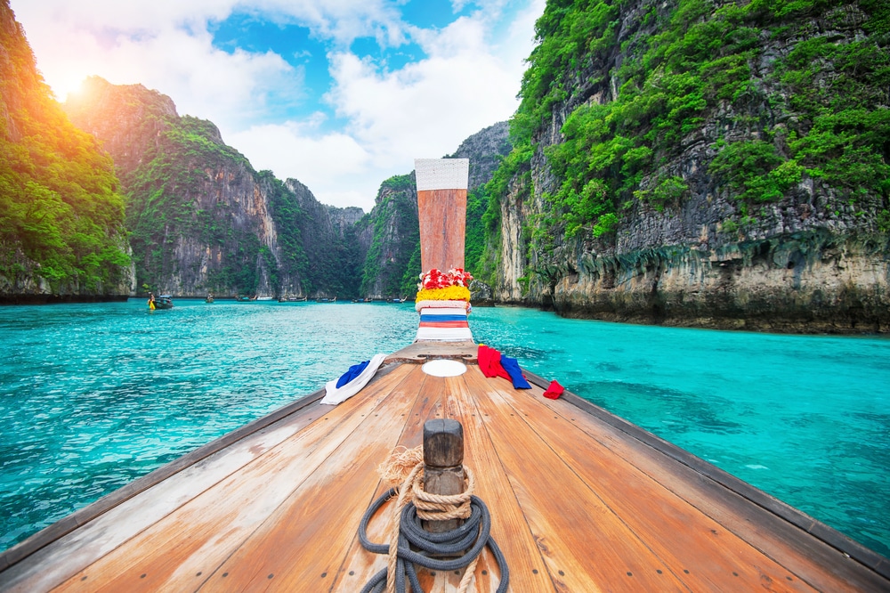 เรือไม้ลำหนึ่งแล่นผ่านน้ำสีฟ้าใสราวเทอร์ควอยซ์ระหว่างหน้าผาสูงชันสีเขียวภายใต้ท้องฟ้าที่มีเมฆบางส่วนและแสงแดด—ภาพที่สมบูรณ์แบบสำหรับการเที่ยวกระบี่.