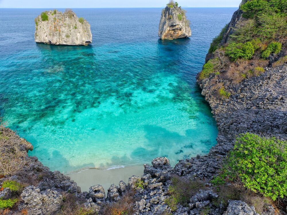 ชายหาดทรายเล็ก ๆ ที่มีน้ำทะเลใสสีเทอร์ควอยซ์ล้อมรอบด้วยหน้าผาหิน พร้อมด้วยหินขนาดใหญ่สองก้อนในทะเลใกล้ ๆ เหมาะสำหรับผู้ที่มองหาสถานที่ท่องเที่ยวในกระบี่หรือวางแผนที่จะเที่ยวในกระบี่.