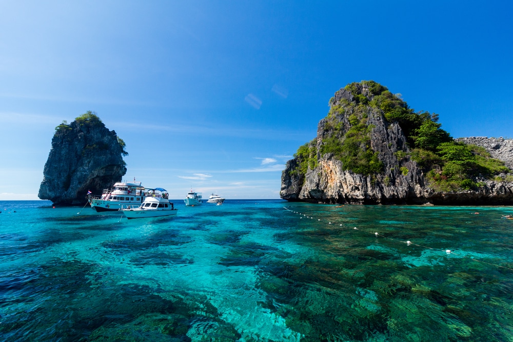 น้ำทะเลใสสีฟ้าที่สามารถมองเห็นปะการังใต้ผิวน้ำได้ มีเรือหลายลำจอดทอดสมออยู่ และมีเกาะหินสองเกาะที่มีพืชพรรณเขียวขจีอยู่ใต้ท้องฟ้าสีฟ้าสดใส—เหมาะสำหรับผู้ที่กำลังมองหาสถานที่ท่องเที่ยวในกระบี่ที่สวยงามท่ามกลางธรรมชาติ.