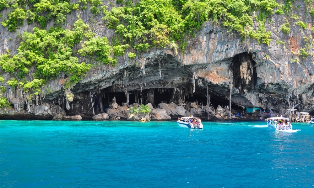 เรือสองลำที่มีนักท่องเที่ยวอยู่ใกล้หน้าผาหินที่มีถ้ำและพืชพรรณเขียวชอุ่ม อยู่ข้างน้ำทะเลใสสีเทอร์ควอยซ์—ฉากที่สมบูรณ์แบบสำหรับผู้ที่ต้องการ เที่ยวกระบี่ หรือค้นหาสมบัติที่ซ่อนอยู่ที่ ที่เที่ยวกระบี่.