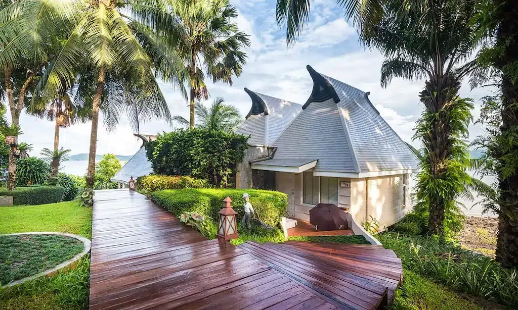 ทางเดินไม้ทอดยาวไปยังบังกะโลสีขาวหลังคาแหลม รายล้อมด้วยต้นปาล์มและพืชพรรณเขียวชอุ่มใต้ท้องฟ้าที่มีเมฆบางส่วน— ที่พักระนอง อันเงียบสงบ เหมาะสำหรับการพักผ่อนครั้งต่อไปของคุณ.