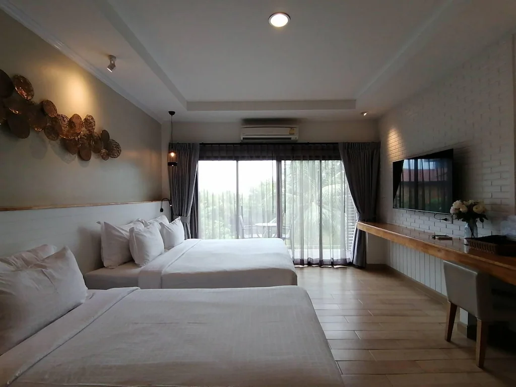 ห้องพักโรงแรมสมัยใหม่พร้อมเตียงคู่สองเตียง โทรทัศน์ติดผนัง โต๊ะทำงานยาว และหน้าต่างบานใหญ่พร้อมผ้าม่านที่มองเห็นระเบียงและพื้นที่สีเขียว—เหมาะสำหรับผู้ที่มองหาที่พักติดทะเลในปราณบุรีราคาไม่แพงสำหรับการพักผ่อนที่ผ่อนคลาย.