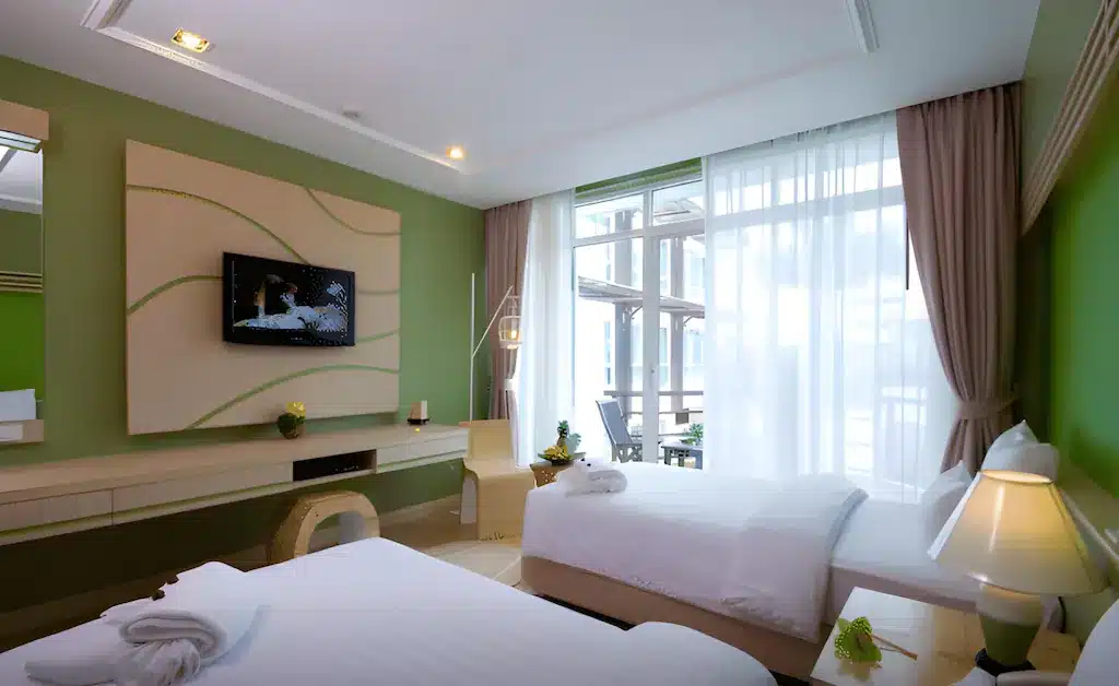 ห้องพักโรงแรมสมัยใหม่พร้อมเตียงเดี่ยวสองเตียง ผนังสีเขียวเป็นจุดเด่น ทีวีติดผนัง โต๊ะทำงาน และหน้าต่าง ที่เที่ยวชุมพร บานใหญ่—เหมาะสำหรับการพักผ่อนหลังจากสำรวจสถานที่ท่องเที่ยวในชุมพรหรือค้นพบสถานที่ท่องเที่ยวใกล้เคียง.