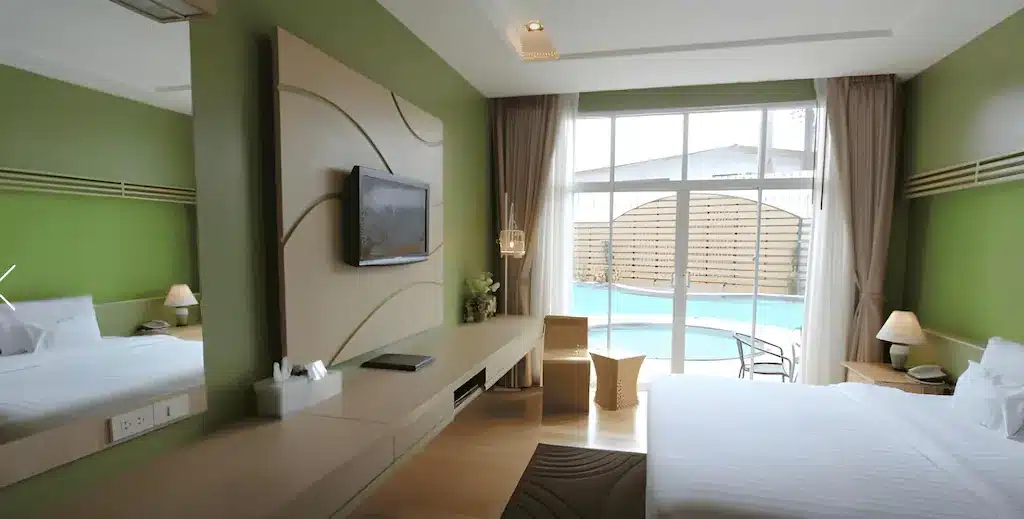 ห้องพักโรงแรมสมัยใหม่พร้อมผนังสีเขียว เตียงคู่ โทรทัศน์ติดผนัง โต๊ะทำงาน และหน้าต่างบานใหญ่ที่มองเห็นสระว่ายน้ำส่วนตัว—เหมาะสำหรับการพักผ่อนหลังจากเที่ยวชมชุมพรหรือสำรวจสถานที่ท่องเที่ยวที่ได้รับความนิยมในชุมพร.