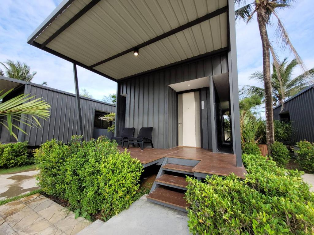 กระท่อมสีดำสมัยใหม่พร้อมเฉลียงที่มีหลังคาคลุม เก้าอี้สีดำสองตัว และบันไดนำขึ้นด้านบน ล้อมรอบด้วยพุ่มไม้สีเขียวและต้นปาล์ม—เหมาะสำหรับการพักผ่อนอย่างมีสไตล์ที่ ที่พักระนอง 