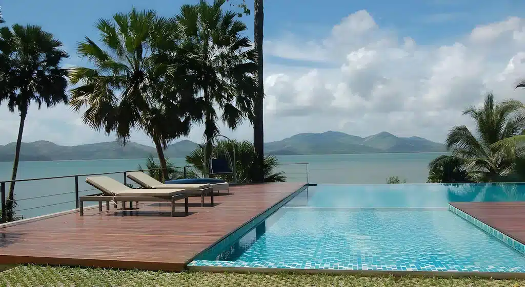 เก้าอี้เลานจ์สองตัววางอยู่บนระเบียงไม้ข้างสระว่ายน้ำแบบอินฟินิตี้ที่มองเห็นน้ำทะเลที่เงียบสงบ ต้นปาล์ม และภูเขาไกลๆ—เหมาะสำหรับผู้ที่ต้องการ ที่พักระนอง ติดทะเลพร้อมวิวที่สวยงามตระการตา.