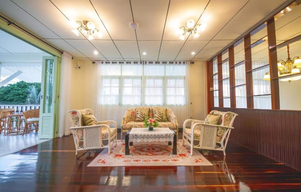 ห้องนั่งเล่นที่อบอุ่นพร้อมชุดโซฟา ที่พักระนอง ลายดอกไม้ โต๊ะกาแฟกระจก พรมลาย และพื้นไม้—เหมาะสำหรับการพักผ่อนที่ ที่พักระนอง—ติดกับพื้นที่รับประทานอาหารที่สว่างสดใสพร้อมประตูเปิดโล่ง.