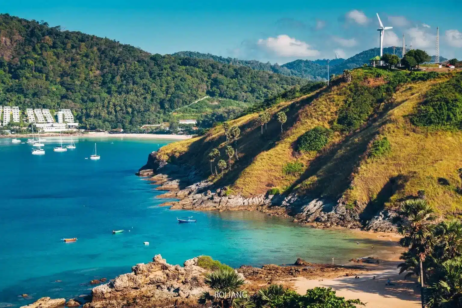 ทิวทัศน์ชายฝั่งที่มีน้ำทะเลสีคราม เรือใบ ชายหาดทรายขาว ภูเขาเขียวขจี และกังหันลมบนยอดเขาภายใต้ท้องฟ้าแจ่มใส—ถ่ายทอดเสน่ห์ของ ที่เที่ยวป่าตอง ได้อย่างสมบูรณ์แบบ.