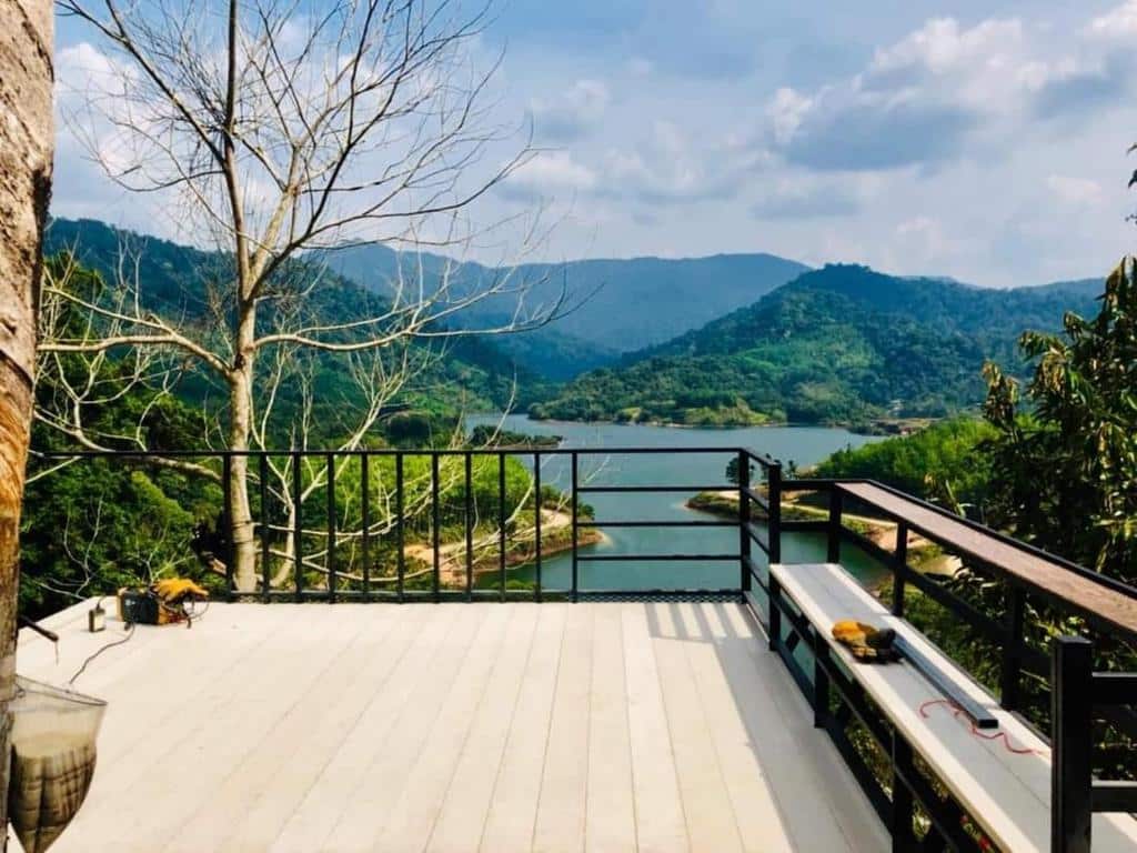 ระเบียง ที่พักระนอง ที่มีพื้นสีขาวทอดมองออกไปยังทะเลสาบซึ่งล้อมรอบด้วยเนินเขาและภูเขาสีเขียวภายใต้ท้องฟ้าที่มีเมฆบางส่วน—ทัศนียภาพอันเงียบสงบที่มักพบได้ที่ ที่พักระนองในเมือง. ต้นไม้ไร้ใบตั้งอยู่ทางซ้ายมือ.