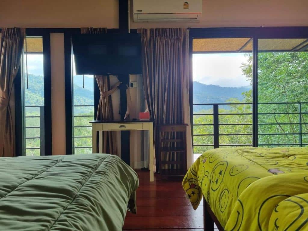 ห้องพักที่ โรงแรมระนอง พร้อมเตียงสองเตียง โต๊ะทำงาน โทรทัศน์ติดผนัง และหน้าต่างบานใหญ่ที่มองเห็นวิวภูเขาและป่าไม้ที่สวยงาม—ตัวเลือกที่สมบูรณ์แบบสำหรับการพักผ่อนในระนองที่มอบความสะดวกสบายและบรรยากาศธรรมชาติ.