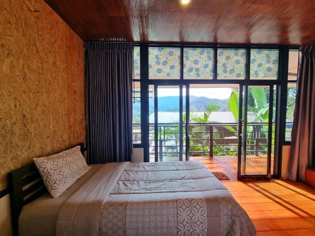 ห้องนอนที่ ที่พักระนอง สว่างไสวด้วยแสงแดด มีเตียงคู่ ผ้าปูที่นอนลายสวย และประตูบานใหญ่กระจกใสที่เปิดออกสู่ระเบียงพร้อมวิวภูเขาและต้นไม้เขียวขจี—เหมาะสำหรับการเข้าพักที่ โรงแรมระนอง ที่แสนอบอุ่นของคุณ.