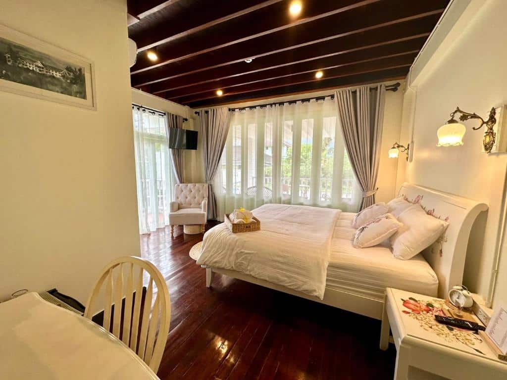 ห้องนอนสว่างไสวใน ที่พักระนองติดทะเล ประกอบด้วยเตียงขนาดใหญ่, พื้น ที่พักระนอง ไม้, ผนังสีขาว, เก้าอี้มีที่วางแขน, โต๊ะทำงาน, และประตูระเบียงพร้อมม่านบางเบา.